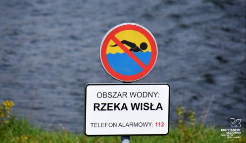 Znak zakazujący kąpieli z tabliczką informacyjną, na której jest tekst: obszar wodny – rzeka Wisła, telefon alarmowy 112. W tle nieostre fale płynącej rzeki.  