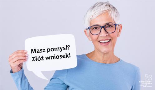 Na zdjęciu uśmiechnięta kobieta – seniorka (krótkie siwe włosy, czarne okulary, ubrana jest w niebieską bluzkę), kobieta trzyma tabliczkę, z napisem: w pierwszym wierszu: Masz pomysł? w drugim wierszu: Złóż wniosek!