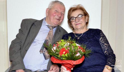 Na fotelach siedzi opisane w tekście małżeństwo. Mężczyzna w szarym garniturze po lewej, kobieta w niebieskiej sukience po prawej – głowy mają zwrócone ku sobie, kobieta trzyma w ręku bukiet kwiatów.