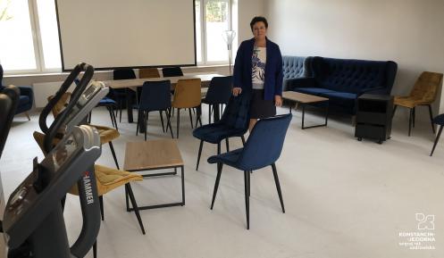 Kobieta stoi w pomieszczeniu z krzesłami i stołami, w tle ekran do projektora.