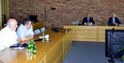 Sala konferencyjna urzędu. Na pierwszym planie dwaj mężczyźni siedzący przy jasnobrązowym stole, na którym stoi mikrofon,  butelki z wodą i szklanki, jeden z nich mówi do mikrofonu. Dalej pod ścianą z czerwonej cegły stoi jasnobrązowy stół, za którym siedzą dwaj mężczyźni – burmistrz i jego zastępca. 