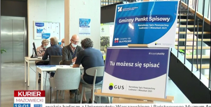 Przestronny korytarz, w którym ustawione są dwa stoliki. Przy nich siedzą ludzie i rozmawiają. Po prawej stronie na sztaludze malarskiej wisi plakat z informacją: Gminny Punkt Spisowy, tu możesz się spisać. 