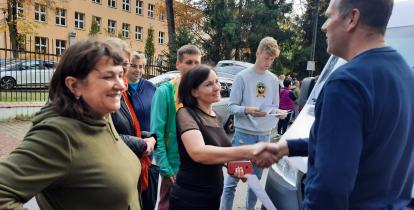 Kilka osób, jedna z nich, kobieta, podaje rękę mężczyźnie w geście podziękowania.