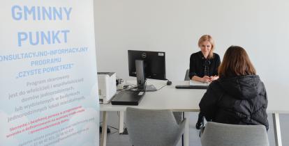 Dwie osoby (kobiety) siedzą przy biurku, na którym stoi monitor komputerowy.