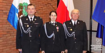 Sala, obok siebie w rzędzie stoi trzech strażaków (w tym jedna kobieta). Ubrani są w stroje galowe.