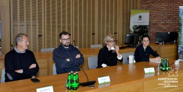 Sala konferencyjna. Cztery osoby siedzą na krzesłach przy drewnianym stole. Dwóch mężczyzn i dwie kobiety. Przed nimi stoją dwa mikrofony, butelki z wodą.