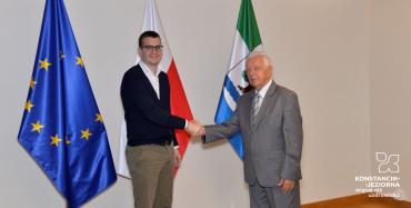 Sala konferencyjna. Mężczyzna w średnim wieku oraz młody mężczyzna stoją obok siebie. Ściskają sobie prawe dłonie. W tle biała ściana oraz trzy flagi: flaga Konstancina-Jeziorny, flaga Polski oraz flaga Unii Europejskiej.