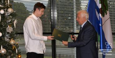 Dwie osoby stoją na tle choinki (nastolatek i mężczyzna). Mężczyzna z prawej (burmistrz) przekazuje młodemu zawodnikowi list gratulacyjny.