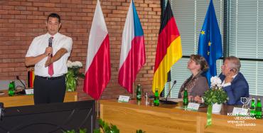 Dwie osoby siedzą jedna stoi i przemawia, w tle flagi 