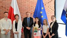 6 osób stoją w sali: 3 dorosłe (2 mężczyzn, 1 kobieta) oraz 3 dziewczynki wieku szkolnym. Jedna dziewczynka trzyma w rękach książkę, którą napisali uczniowie. W tle widać ceglana ścianę. 