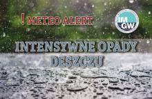 Na górze czerwony napis meteo alert poprzedzony wykrzyknikiem. Obok niego logo Instytutu Meteorologii i Gospodarki Wodnej - niebieski okrąg z białymi literami IMGW, poniżej szary napis intensywne opady deszczu. W tle widać krople deszczu, które spadają na ulice. 