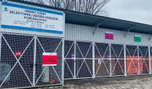 a zdjęciu metalowa podłużna wiata. Pod nią stoi sześć dużych kontenerów budowlanych. Na wiacie powieszony jest duży baner z napisem: Gminny Punkt Selektywnej Zbiórki Odpadów Komunalnych.
