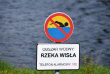 Znak zakazujący kąpieli z tabliczką informacyjną, na której jest tekst: obszar wodny – rzeka Wisła, telefon alarmowy 112. W tle nieostre fale płynącej rzeki.  