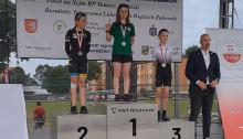 Podium. W środku stoi dziewczyna ze złotym medalem na szyi i zielonej bluzce. Po prawej i lewej jej stronie inne dziewczyny również z medalami na szyi (srebro i brąz).  Obok podium stoi młody mężczyzna w marynarce, dżinsach i białej koszuli.