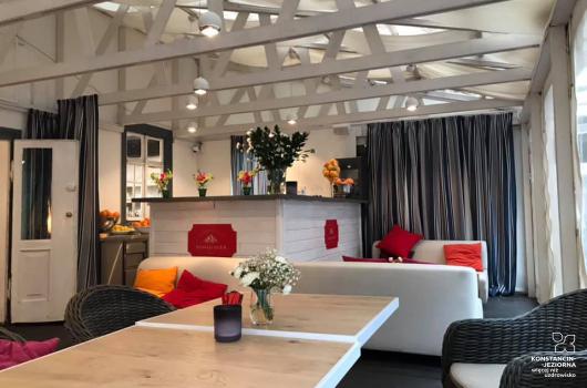 Sala restauracyjna z zadaszeniem belkowanym, z widocznymi stolikami fotelami i sofami, pośrodku bar z szyldem 