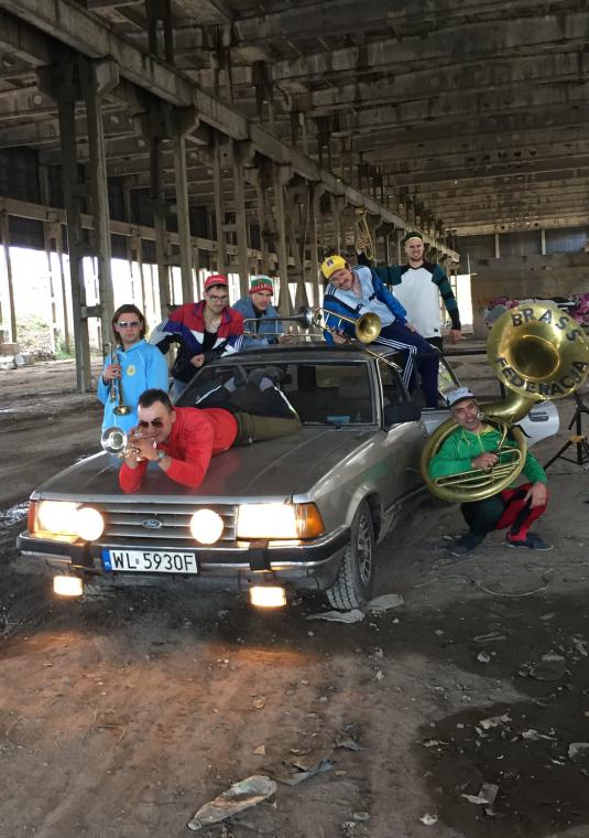 Grupa muzyków z instrumentami dętymi, pośrodku samochód osobowy, dwóch muzyków leży na samochodzie, nad nimi konstrukcja mostu.